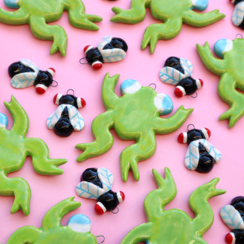 Ceramic Frogs and Flies Holiday Ornaments spread out across a Pink Background made by Eva Stalinski 2022