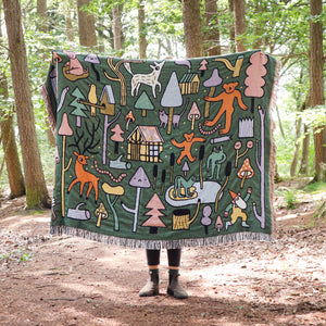 Picture of Eva Stalinski holding  a woven Fairytale Forest Throw Blanket against a backdrop of trees.